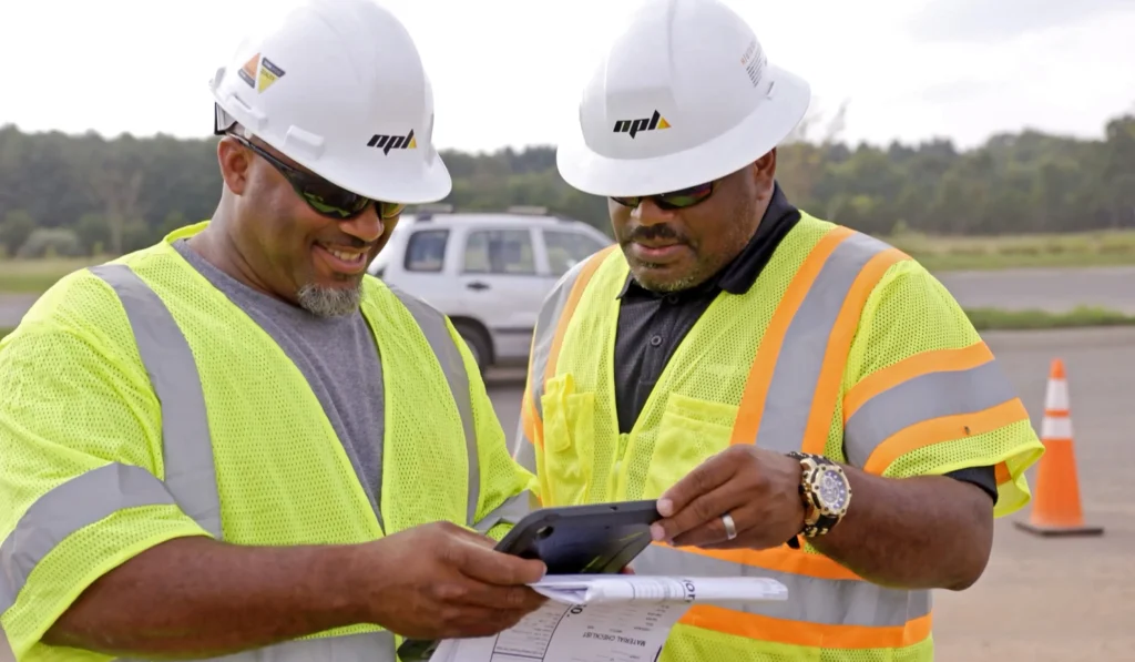 Two workers in hard hats smiling and looking over a tablet